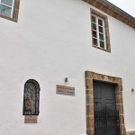 Photograph of the facade of the International Center for the Reception of the Pilgrim. You can see the white facade with the stone door and window frames. To the left of the entrance there is a stone figure of Our Lady of Remedies embedded in the wall with a small fence.