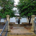 Photograph of the exterior stone stairs ascent with black metal handrails. In front of the stairs there is a circular stone fountain with 4 camelliums around it. In the background is a white entrance door to the building.