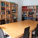 Fotografía detalle de la Biblioteca de la Oficina del Peregrino en donde aparecen librerías llenas de libros y una mesa vacía de madera rectangular con 8 sillas a su alrededor.